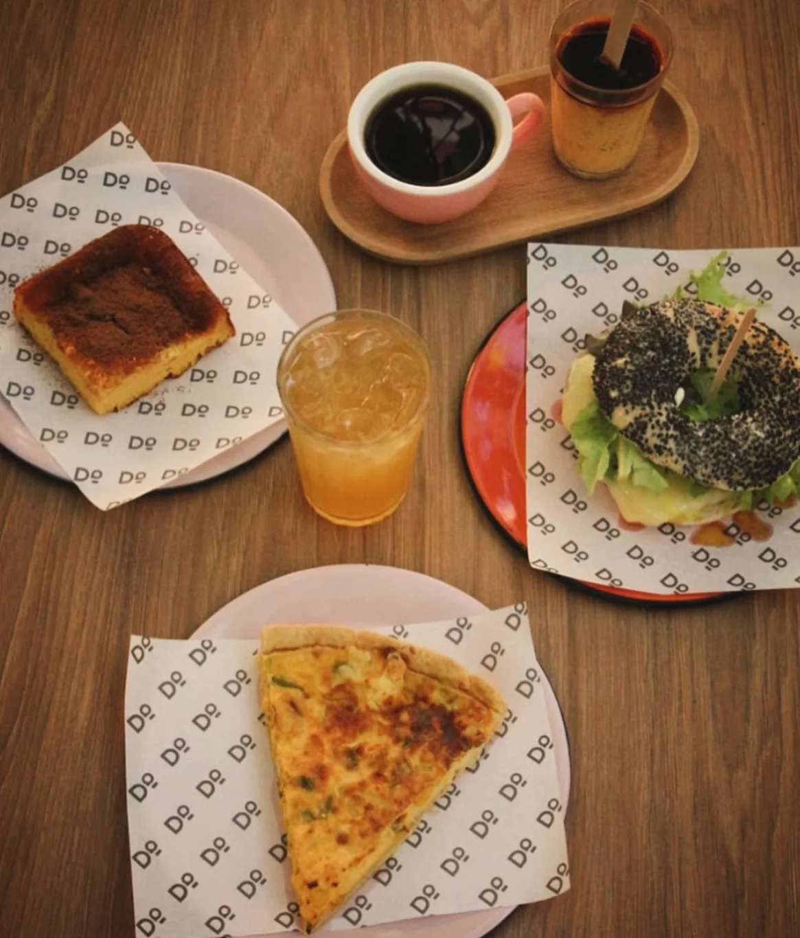Mesa vista de cima com quiche, bagel, suco e café sobre papel Dº